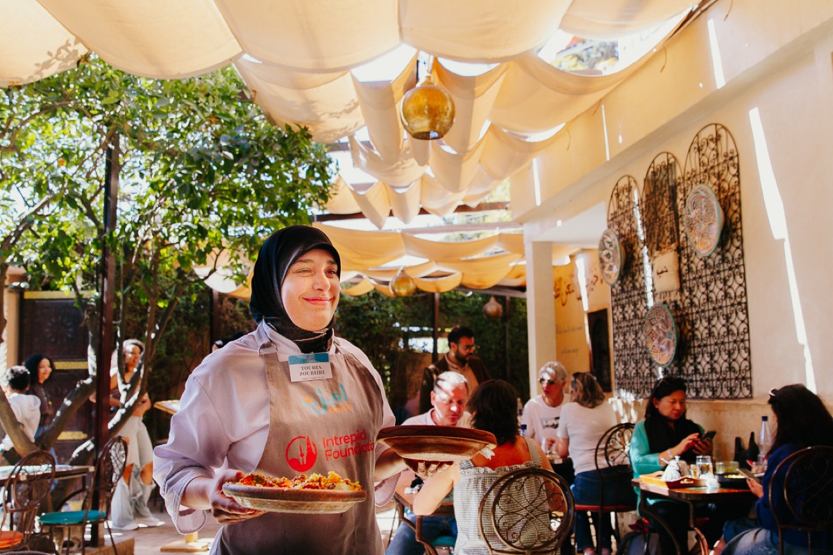 An Amal trainee in apron serving guests on a sunny garden terrace